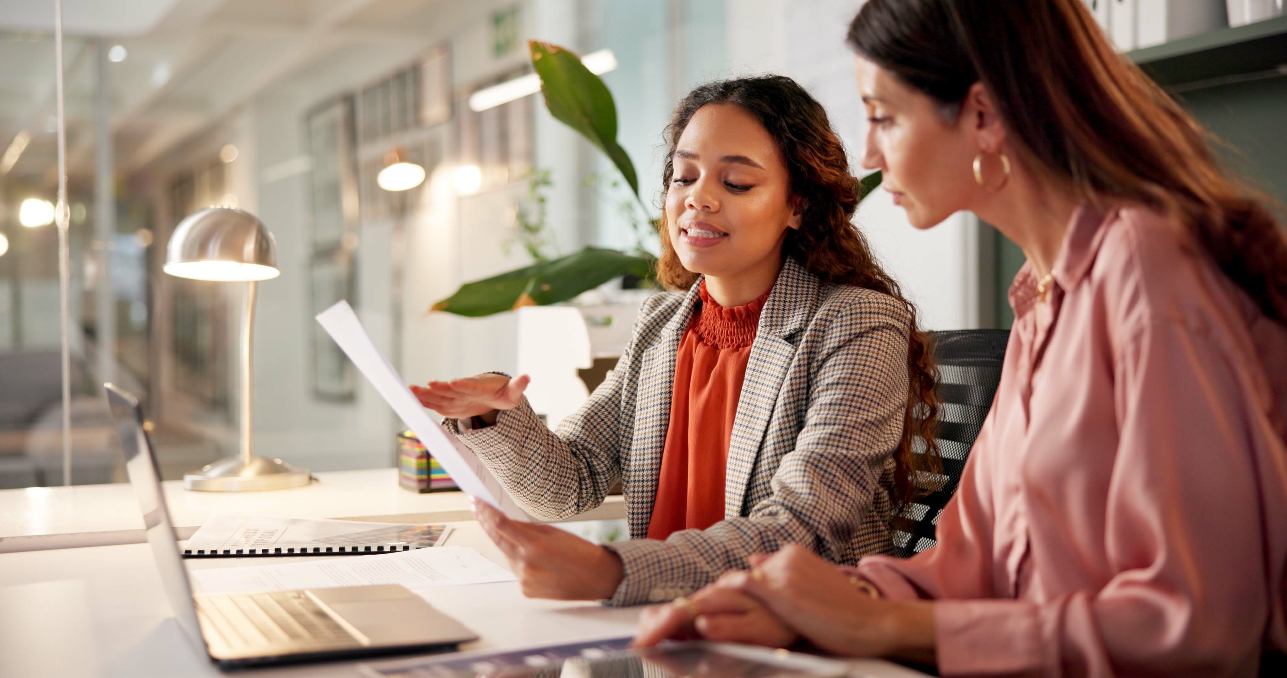 Two women meeting at a table with laptops and papers, symbolizing personal financial coaching or planning support